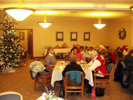 A wide view of the dining room.  Shirley, Dottie, Gloria, Emelia, and June sit at a table with other tables behind them and next to them. The Christmas tree and table with presents are in the background.