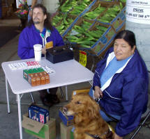 Terry and Viola, with her dog guide Alberta, sitting at a table selling candy